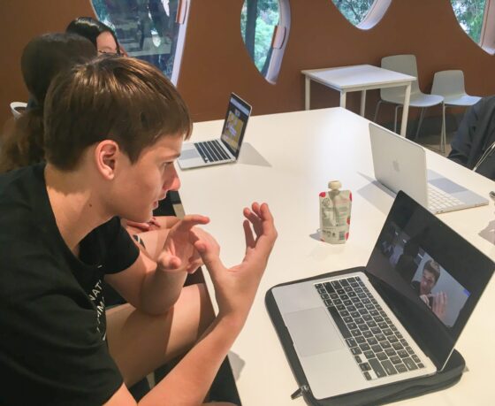 A young Japanese secondary school student sits at a table, using sign language while video chatting on a laptop as part of an online exchange programme. Others with laptops sit nearby, and the background features round windows and modern furniture.