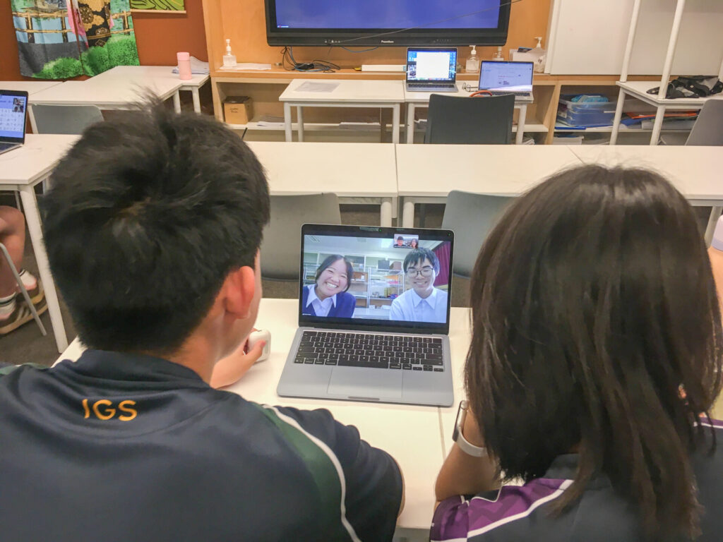 Two Japanese secondary school students sit at a desk, video calling two other students on a laptop as part of an online exchange programme in a classroom with empty chairs, desks, and laptops in the background.