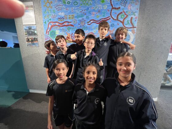 A group of nine smiling IGS Decathletes in black uniforms pose in front of a colourful, abstract mural with blue, red, and green patterns on a wall. One child at the back holds up a peace sign, celebrating their recent State competition.