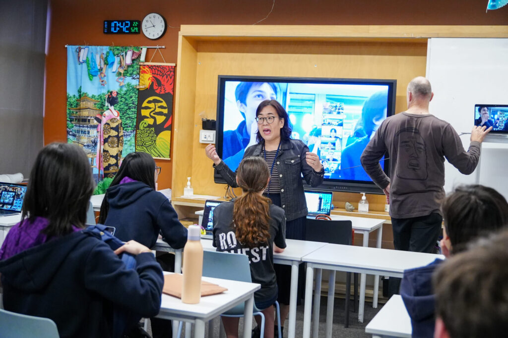 A teacher speaks to Japanese secondary school students in a classroom with a large screen showing an online exchange. Pupils sit at desks with tablets while another adult writes on a whiteboard. Colourful art decorates the room.