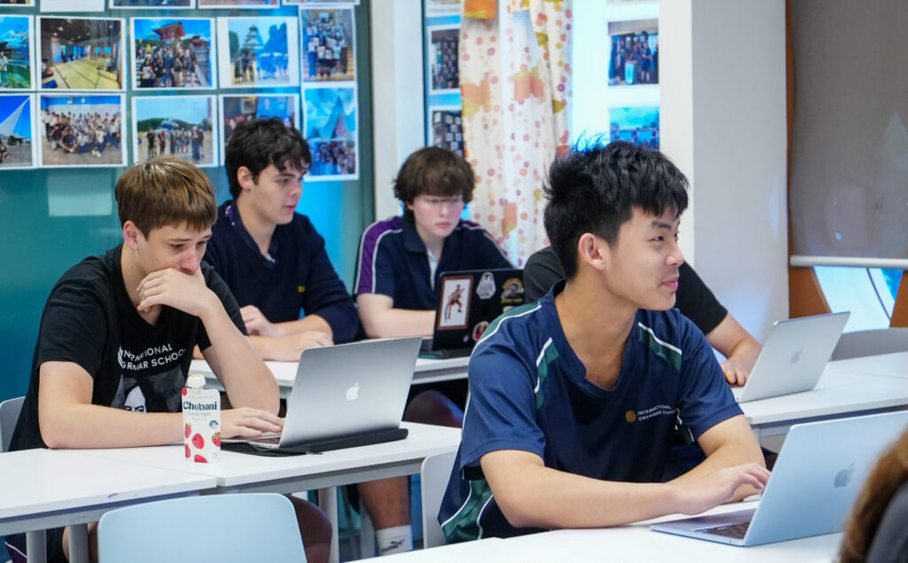 A group of Japanese secondary school students sit at desks in a classroom, each working on a laptop—possibly taking part in an online exchange programme. One student sips from a carton of juice as colourful photos decorate the wall behind them.