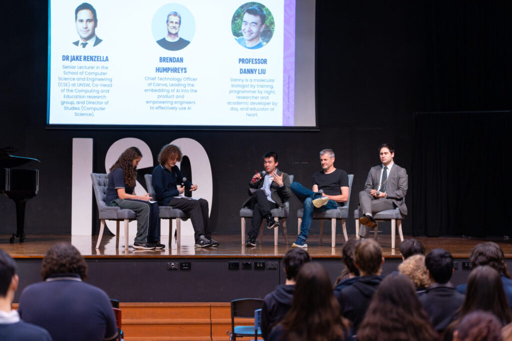 A panel of four speakers from IGS sits on stage in front of an audience during the Day of AI Australia. Behind them, a screen displays their photos, names, and titles. One person speaks into a microphone while others listen attentively to the future-focussed discussion.