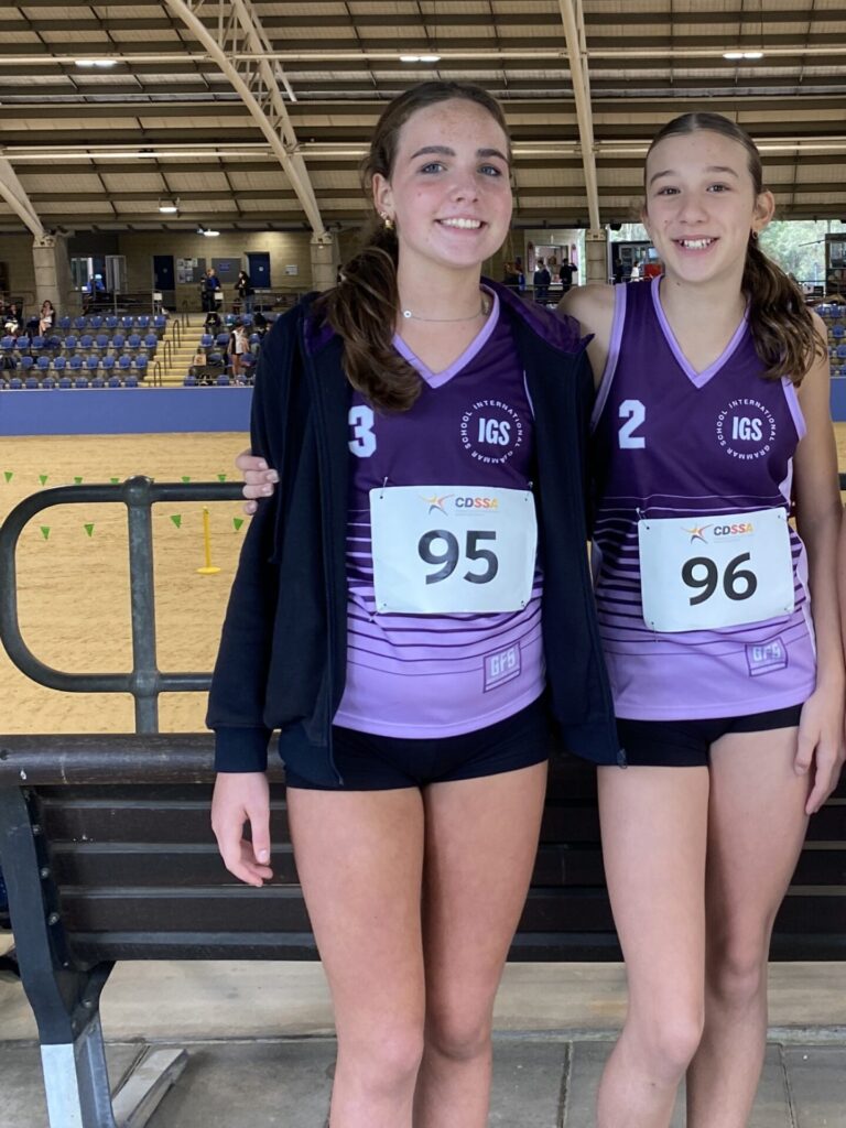 Two teenage girls in matching purple CDSSA cross-country uniforms with race numbers 95 and 96 stand side by side, smiling, in an indoor sports arena with empty seats in the background.