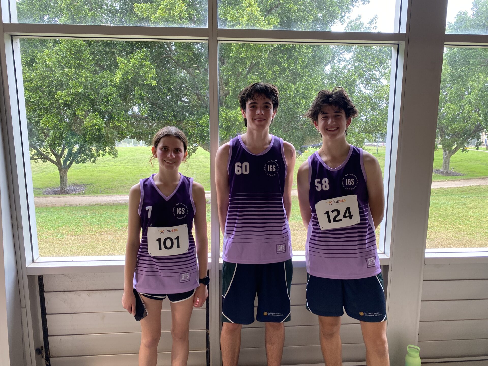 Three teenagers wearing purple CDSSA athletic jerseys and race numbers stand indoors in front of large windows with green trees outside. Smiling and ready for a cross-country event, they appear excited for the race ahead.