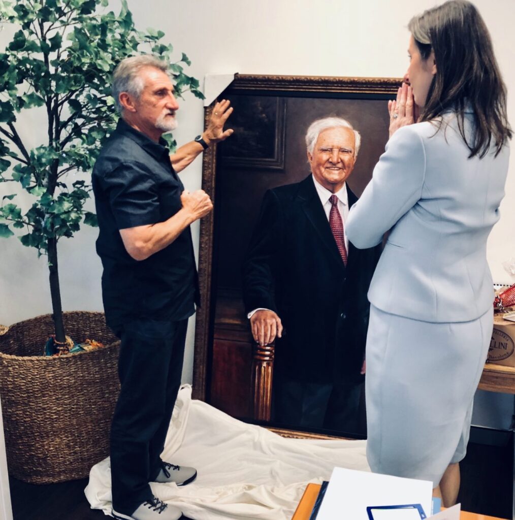 A man and a woman stand near a large framed portrait of Reg St Leon OAM. The man gestures towards the painting during the portrait unveiling, whilst the woman looks at it thoughtfully. A potted tree stands nearby.