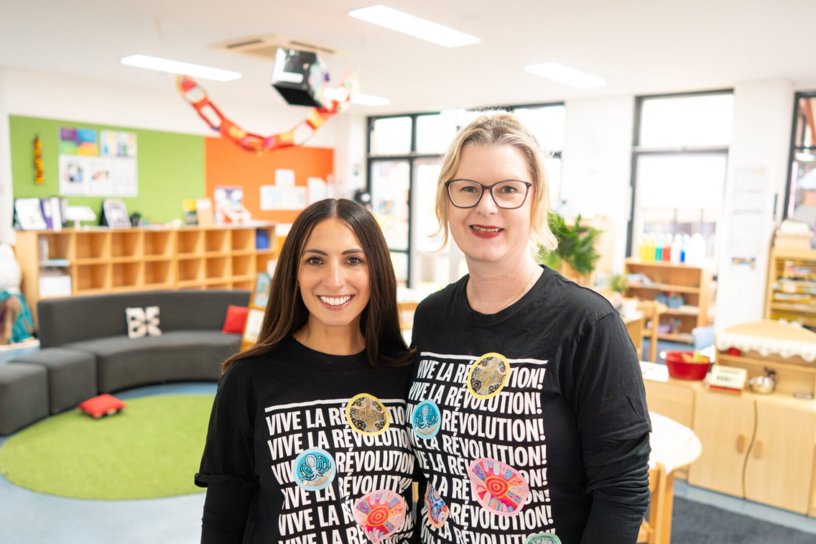 Two women wearing matching Vive La Révolution! shirts smile whilst standing in a brightly lit, colourful Early Learning classroom with shelves, books, and a green carpet area in the background.