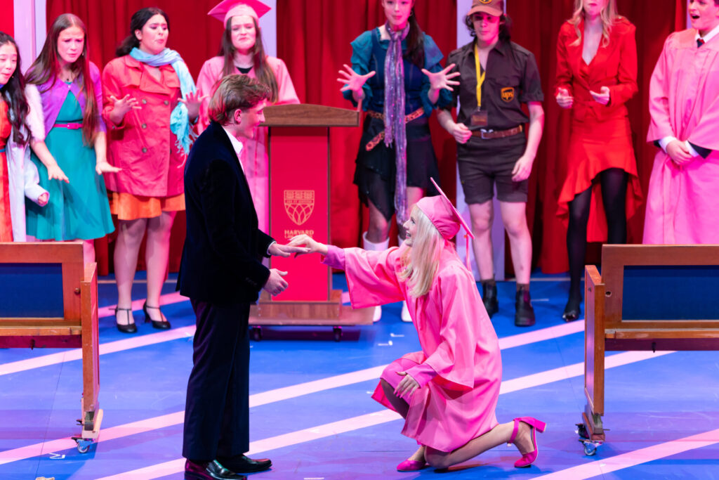 A woman in a pink graduation gown kneels and holds hands with a man in a dinner jacket on stage, as a group of people in colourful costumes watch in the background.