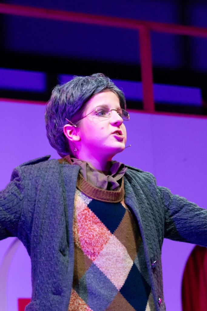 A person on stage in costume with a short wig, glasses, a microphone headset, a textured jacket, and an argyle jumper, stands under purple lighting, speaking or singing with arms raised.