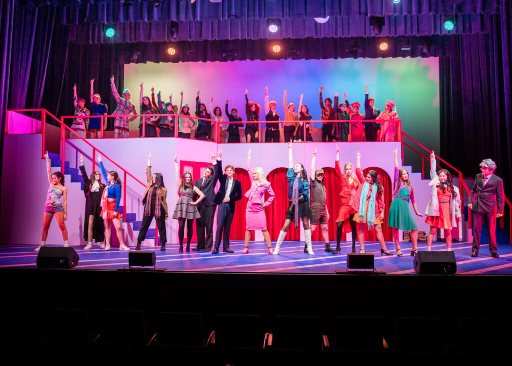 A large group of performers in colourful costumes stand on a brightly lit stage, striking dramatic poses with one arm raised. The set features stairs and arches, with vibrant lighting and a cheering ensemble in the background.