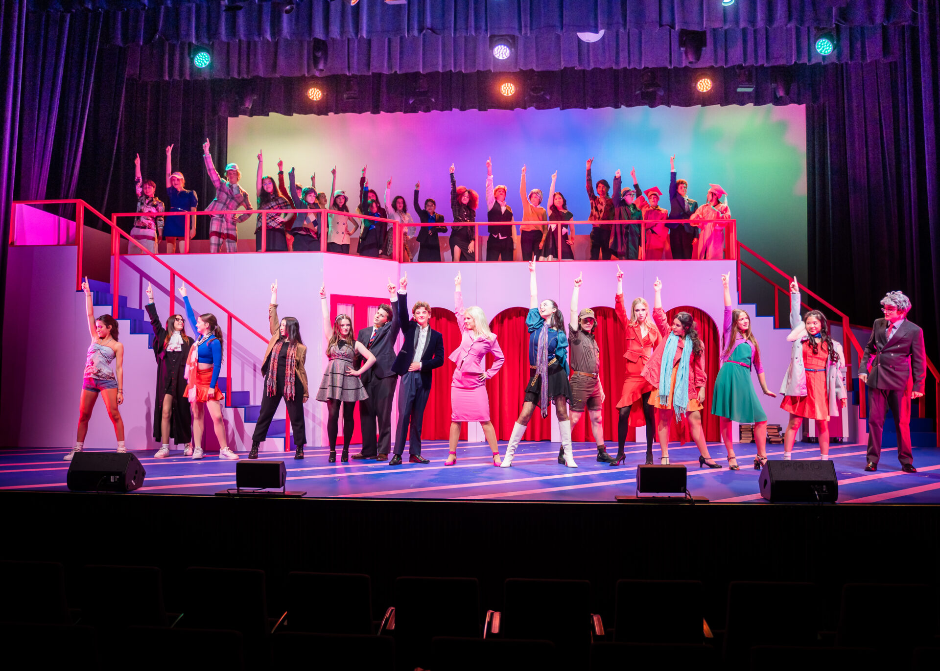 A large group of performers in colourful costumes stand on a brightly lit stage, striking dramatic poses with one arm raised. The set features stairs and arches, with vibrant lighting and a cheering ensemble in the background.