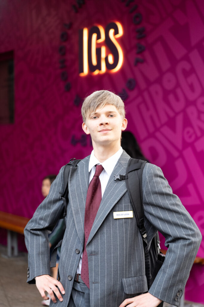 A young person in a pinstripe suit and maroon tie stands confidently with hands on hips, embodying archives in action. They wear a rucksack and name badge, framed by a pink wall with bold lettering and the illuminated acronym IGS above.