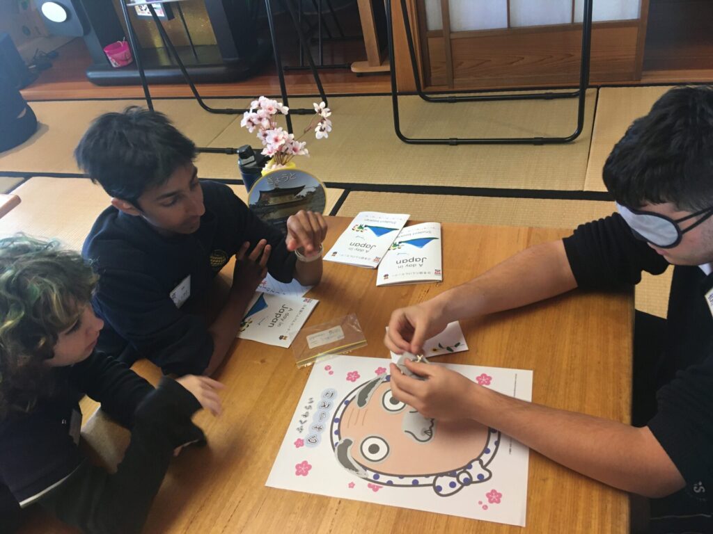 Three young people sit around a low wooden table, enjoying an authentic game inspired by Japanese culture, where one, blindfolded, tries to place facial features on a cartoon face. Leaflets and small decorative items are also on the table.