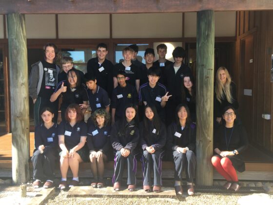 A group of students and two adults, dressed in dark uniforms, are posing for a photo on the veranda of a wooden building, reflecting authentic Japanese culture. Many are smiling at the camera.