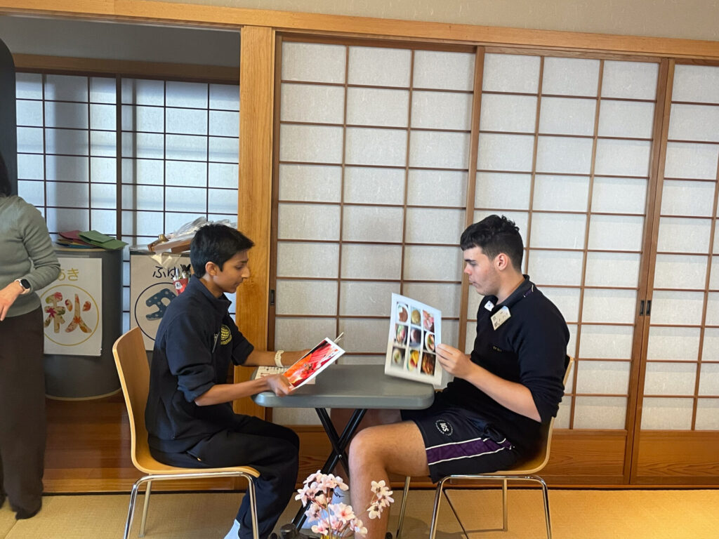 Two people sit opposite each other at a small table, each holding a menu. They appear to be in an authentic Japanese-style room with sliding shoji doors and cherry blossom decorations, reflecting the essence of Japanese culture.