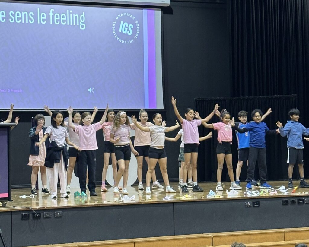 A group of students stands on stage in a row with their arms raised, celebrating learning as they perform in front of a screen displaying “IGS” and text in French. The students wear casual sportswear and appear to be in mid-performance.
