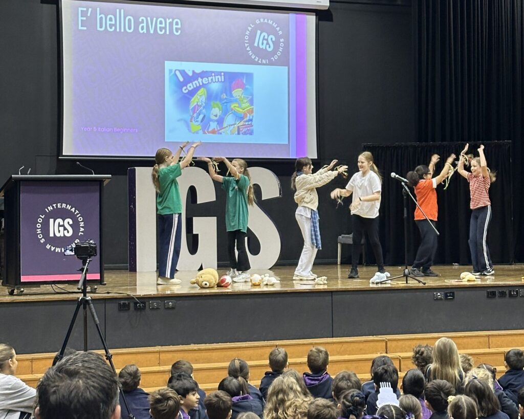 A group of children perform on stage in front of an audience, with a large IGS sign, a lectern, and a screen behind them displaying “E’ bello avere” and an illustration labelled “Canterini,” celebrating languages and learning together.