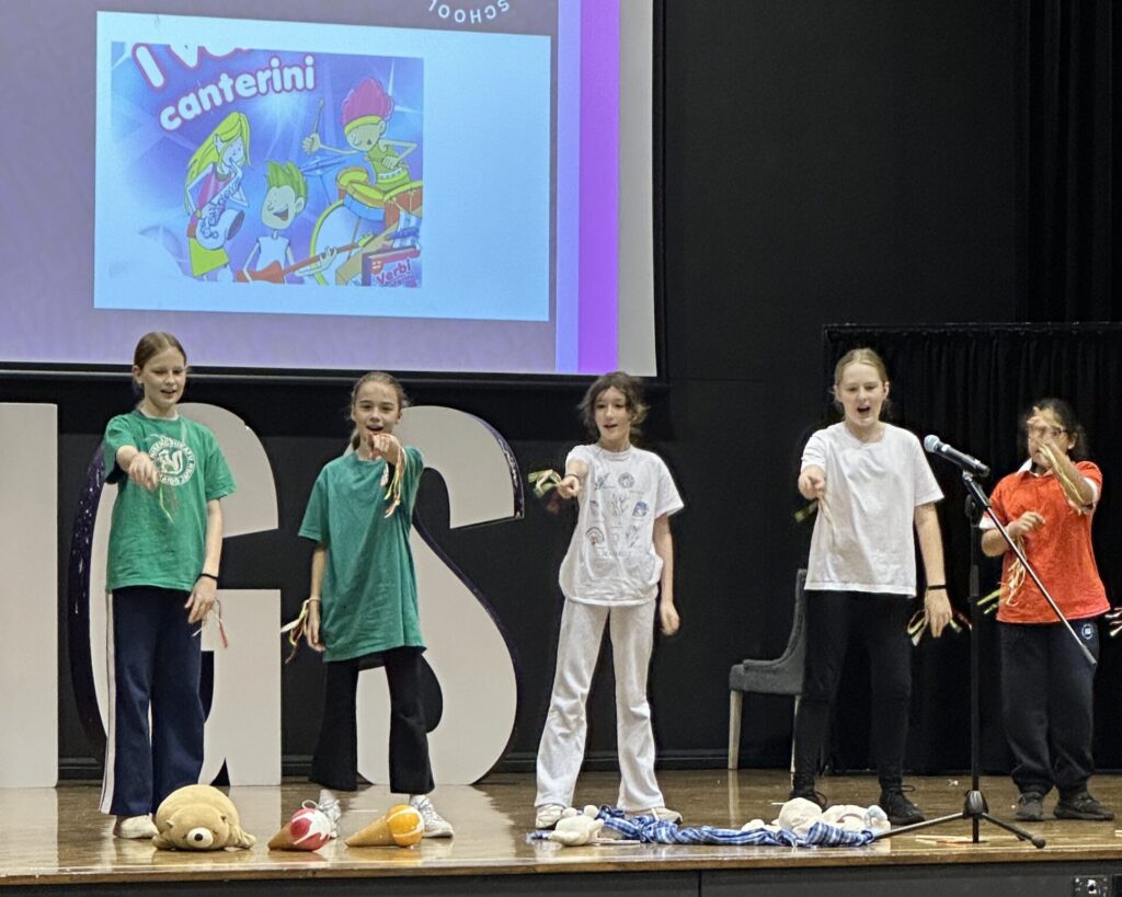 Five children stand on a stage, wearing casual clothes, performing with props in front of a large IGGS sign. A projected cartoon image appears behind them, and soft toys rest by their feet as they celebrate learning together.