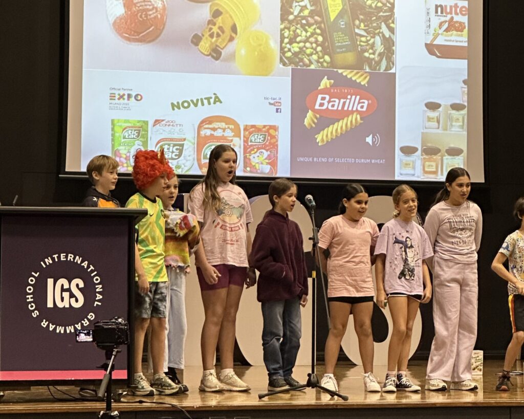 A group of children stands on a stage in front of a lectern labelled IGS and a large screen displaying Italian food brands, celebrating learning languages together. The kids face the audience, some in colourful outfits.