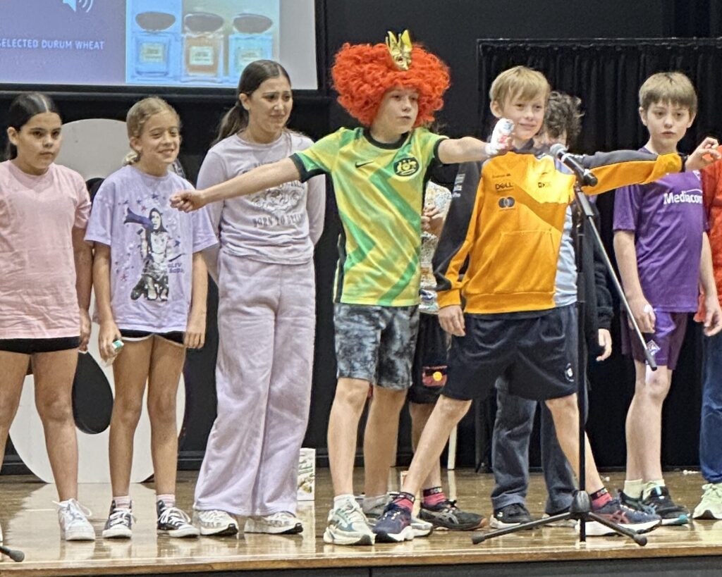 A group of children stand on stage; one boy in a green football shirt and red clown wig gestures widely while others in shorts and T-shirts look on, Celebrating Learning as a microphone stands before them.