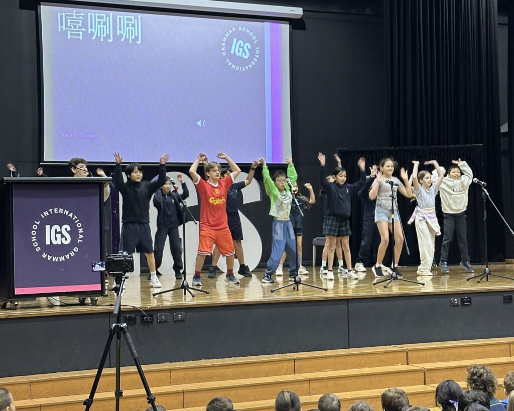 A group of school children energetically perform on stage in front of microphones, with a large screen behind them displaying the IGS logo and Chinese characters, celebrating languages and learning as the audience watches from the seating area.