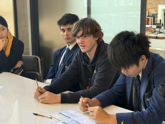 A group of students sit around a table in discussion, holding pencils and papers. One student in casual clothing speaks about the impact of AI whilst others in school uniforms listen attentively.