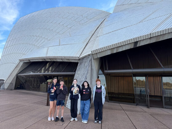 Five young people stand smiling in front of the Sydney Opera House, with its distinctive white tiled sails visible behind them. The group is casually dressed, posing on the building’s broad forecourt.