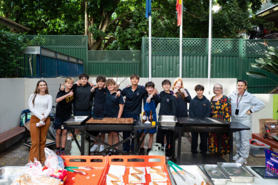A group of students and two adults stand behind barbecue grills, posing for a photo outdoors. Red Earth Ready shirts stand out as they serve sandwiches, with barbecue supplies on the table in front and trees and fences in the background.