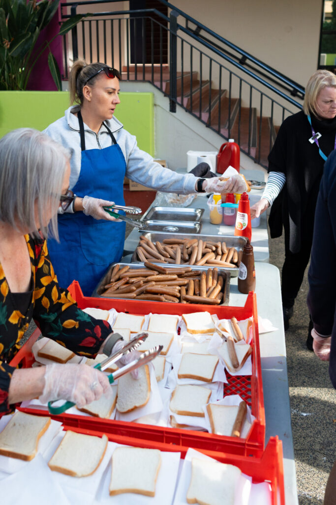 Several people prepare and serve hot dogs at an outdoor table, with trays of sausages and slices of bread. A woman in a blue apron hands out a plate while others assemble food—everyone is Red Earth Ready with condiments visible on the table.