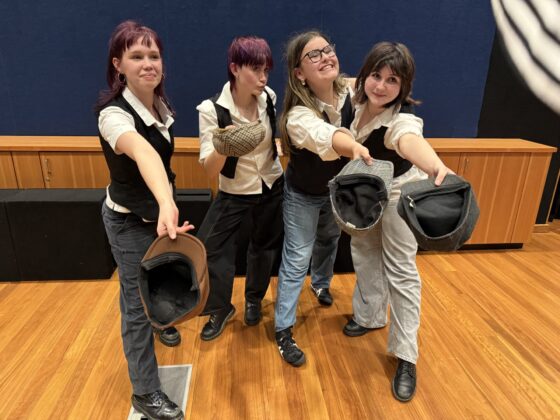Four people wearing black waistcoats and white shirts stand smiling together on a wooden floor with a dark blue wall behind them, holding hats in their outstretched hands—ready to compete as Teams in the Theatresports Semi-Final.