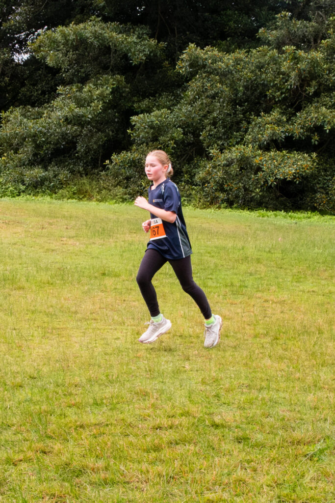 A young girl wearing a race number runs on a grassy field at the Junior School Sports Round-Up, surrounded by green trees. She is dressed in sports clothes and looks focused.