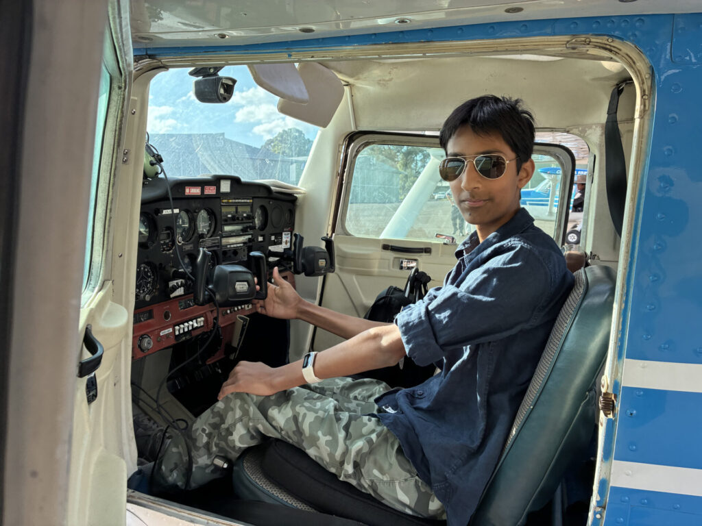 A Year 10 pupil wearing sunglasses and a blue shirt sits in the pilot’s seat of a small aeroplane, holding the controls and looking towards the camera as they take flight with the Royal Aero Club. The plane has a blue and white exterior.