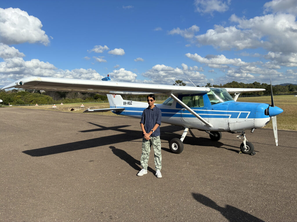 A Year 10 pupil wearing sunglasses and casual clothes stands on an airstrip next to a small blue and white single-engined aeroplane, ready to take flight under a partly cloudy sky.