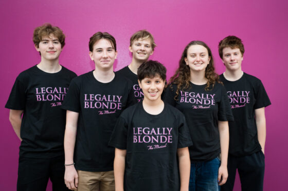 Six smiling teenagers, part of an Extraordinary Team, stand in front of a purple wall, all wearing black Legally Blonde The Musical t-shirts. Five are in the back row and one stands proudly at the front.