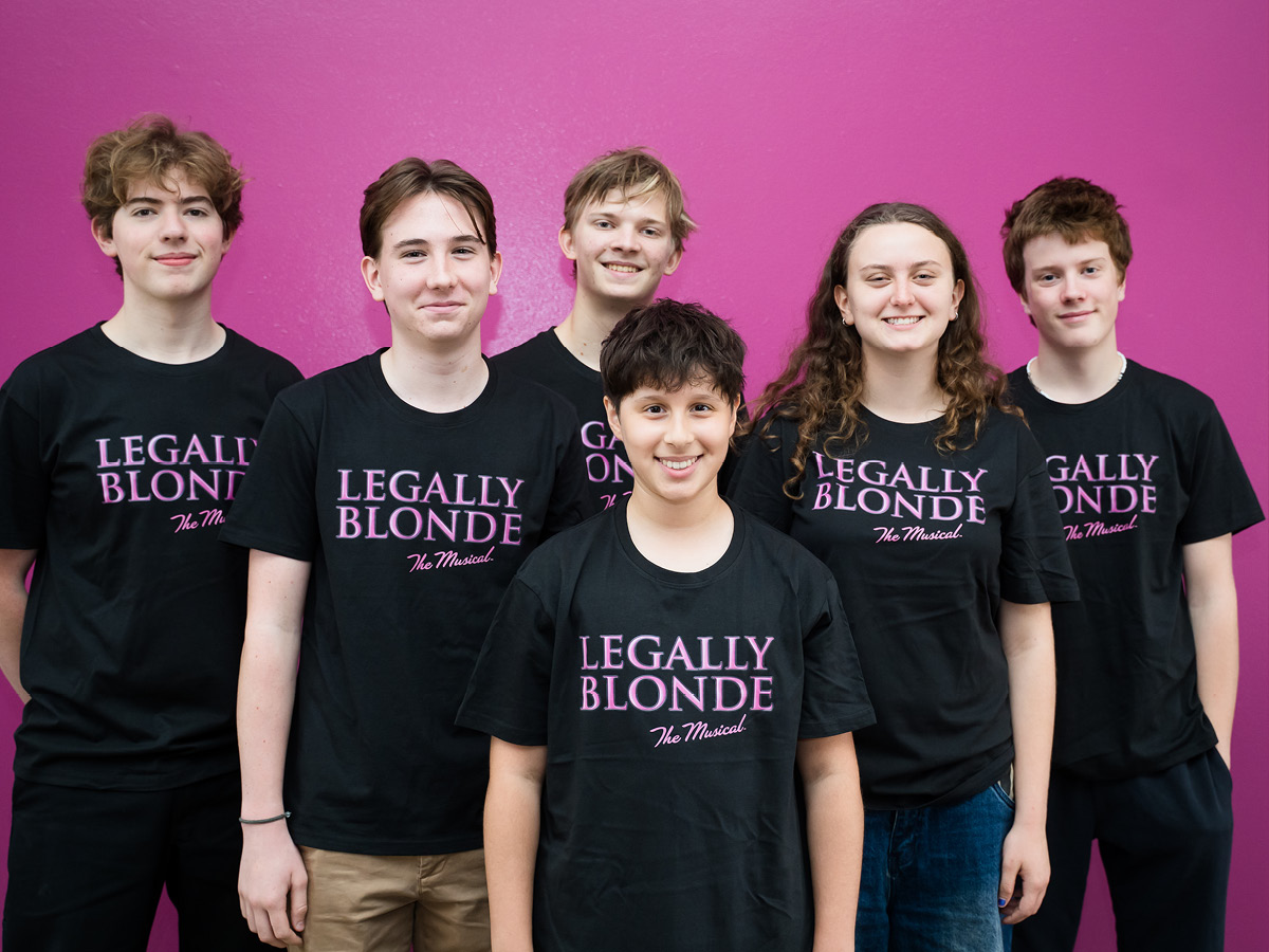 Six teenagers stand in front of a bright pink wall, smiling and wearing matching black Legally Blonde The Musical T-shirts—proud members of the extraordinary team behind the magic of the show.