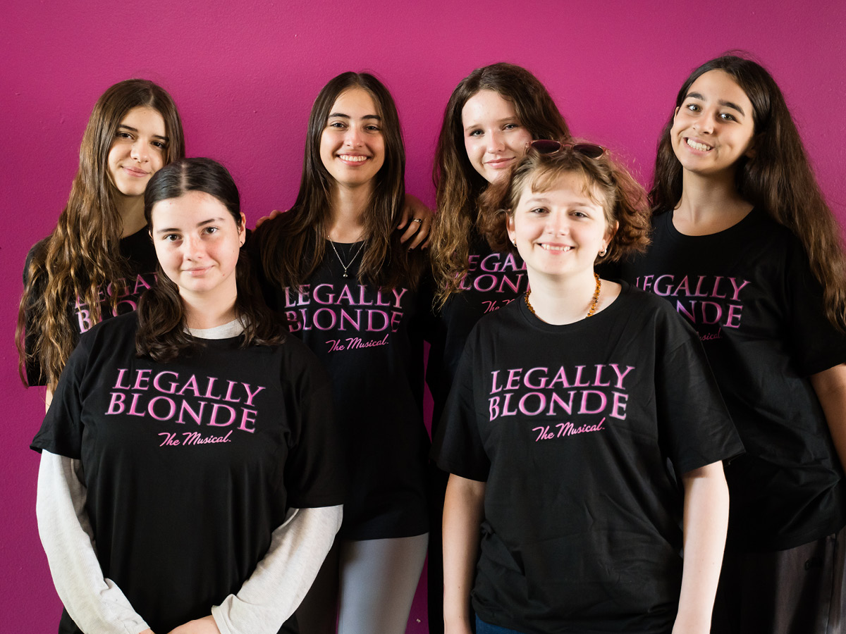 Six smiling young women, the extraordinary team behind the magic, stand in front of a pink wall, proudly wearing black Legally Blonde The Musical T-shirts.