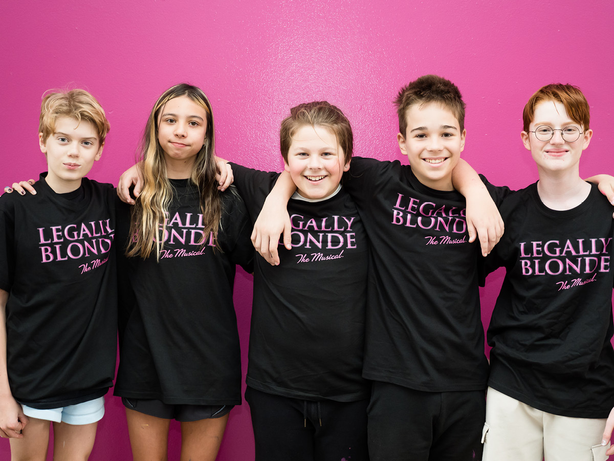 Five smiling kids stand in front of a bright pink wall, arms round each other, wearing matching “Legally Blonde The Musical” t-shirts—behind the magic of this show is an extraordinary team of friends.
