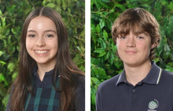 Two teenagers stand in front of a leafy green background, each smiling. The girl on the left has long brown hair and wears a checked dress, while the boy, one of the Year 11 artists in the art programme, sports a navy polo with a school emblem.