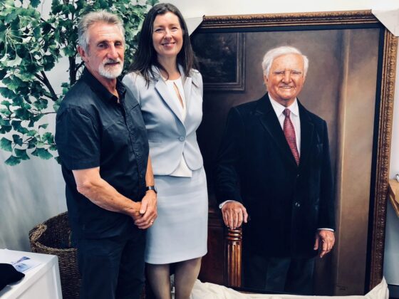 A man and a woman stand smiling next to a large, framed portrait of Reg St Leon, OAM. The portrait unveiling takes place indoors, with a leafy green plant and a wicker basket in the background.