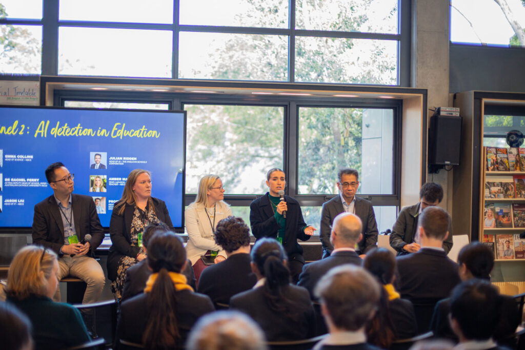 A panel of six speakers sits facing an audience in a modern room, discussing the impact of AI detection in Education as displayed on a screen behind them. Large windows and bookshelves line the background.
