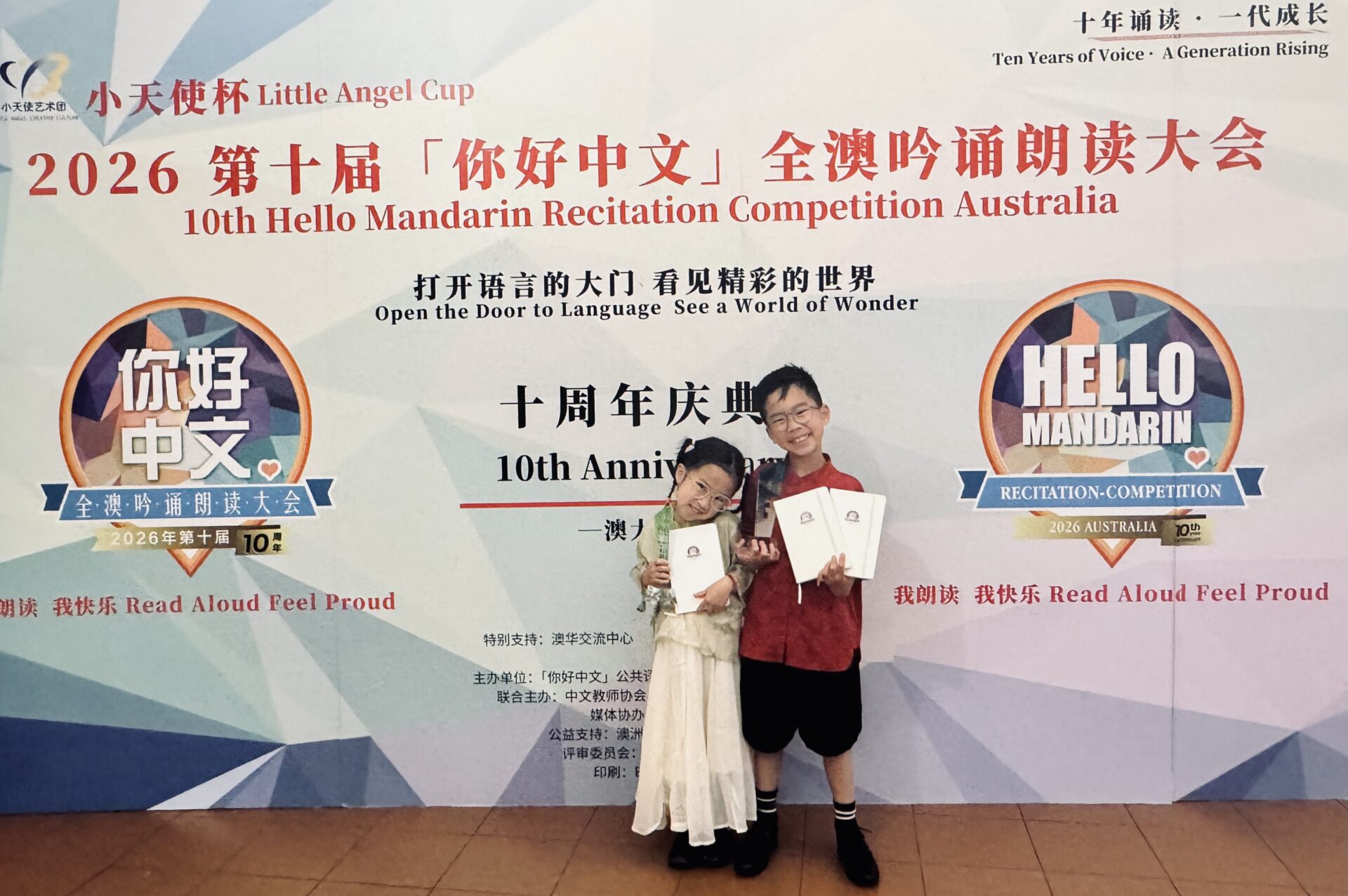 Two smiling children holding certificates stand in front of a colourful backdrop for the 10th Hello Mandarin Recitation Contest, featuring Mandarin Speaking and English text, logos, and event details.