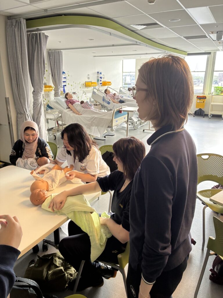 Four students practise infant care using a baby doll and blankets at a table in a bright classroom, exploring future pathways in healthcare. In the background, hospital beds and medical equipment highlight the training setting.