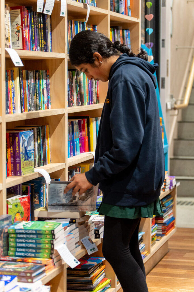 A young person in a dark hoodie stands by bookshelves in a bookshop, discovering new books. Colourful books line the shelves and tables around them as they look at an open book.