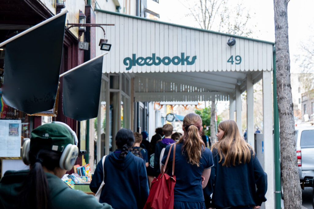 People walk past a white awning with gleebooks written on it. The awning covers a pavement lined with new books and leads to a bookshop entrance at 49 Glebe Point Road. A person with headphones is in the foreground, discovering books along the way.