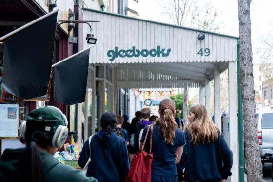 People walk past a white awning with gleebooks written on it. The awning covers a pavement lined with new books and leads to a bookshop entrance at 49 Glebe Point Road. A person with headphones is in the foreground, discovering books along the way.