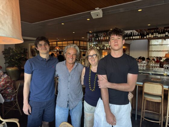 Four people, including Marg Butler, stand together and smile in a stylish restaurant, with a bar and wine bottles in the background. Two young men flank an older man and woman, all dressed in casual, summery outfits.