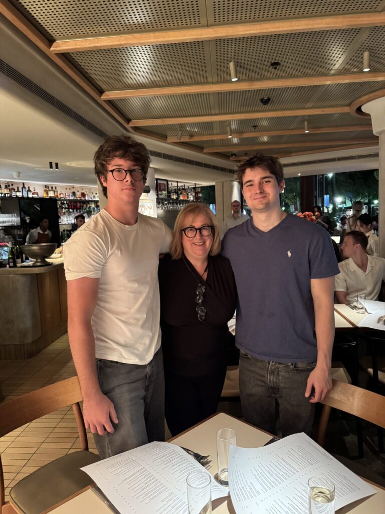 Three people pose together and smile inside a modern restaurant. Marg Butler stands between two young men, with menus and empty glasses on the table in front of them. Other diners and bar shelves are visible in the background.