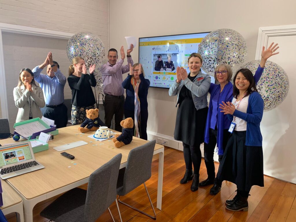 A group of eight people stand smiling and cheering in a bright office room, with large confetti balloons, a screen showing Marg Butler on a video call, and a table with laptops, papers, and teddy bears in the foreground.