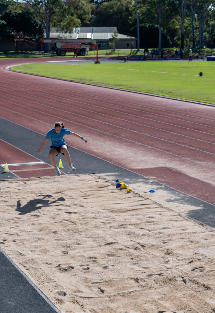 A young athlete in a blue shirt is mid-air during a Colour competition at an outdoor track and field facility, aiming to land in a sandpit marked with coloured cones.