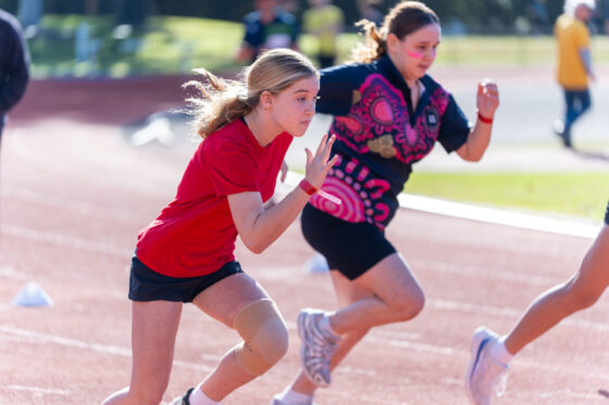 Two young athletes sprint on a track, embodying house spirit. The girl in front wears a red shirt, black shorts, and a knee support. Another runner in black shorts and a patterned top adds colour and competition as they race with focus and energy.