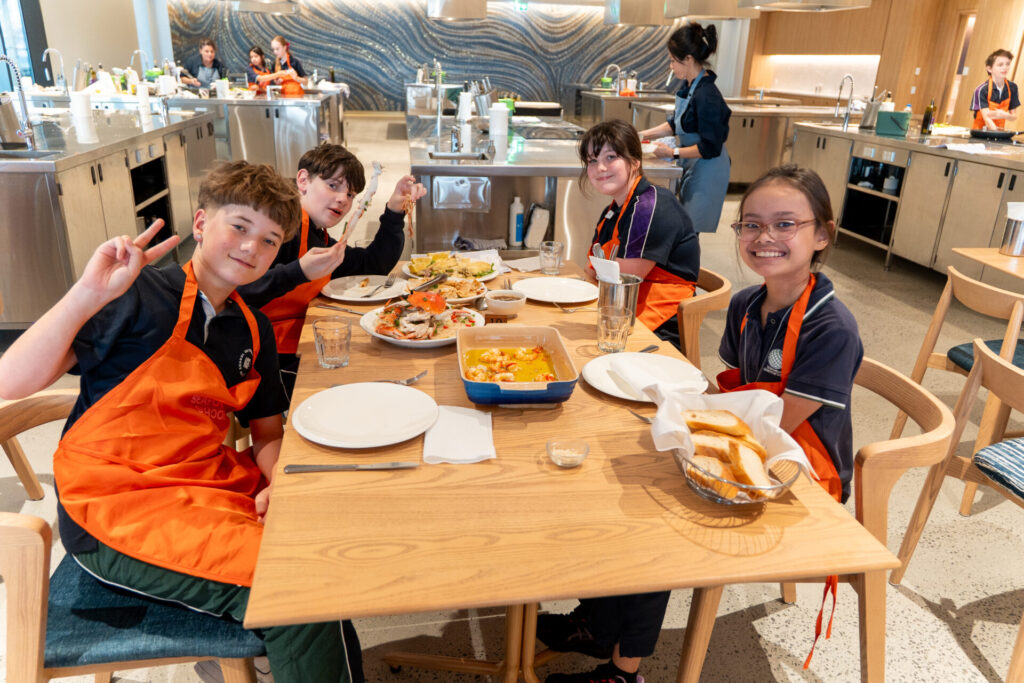 Four smiling secondary school children wearing orange aprons sit around a table in a kitchen classroom at Sydney Fish Market, enjoying a meal together. Various dishes and utensils are on the table, while two adults work in the background.